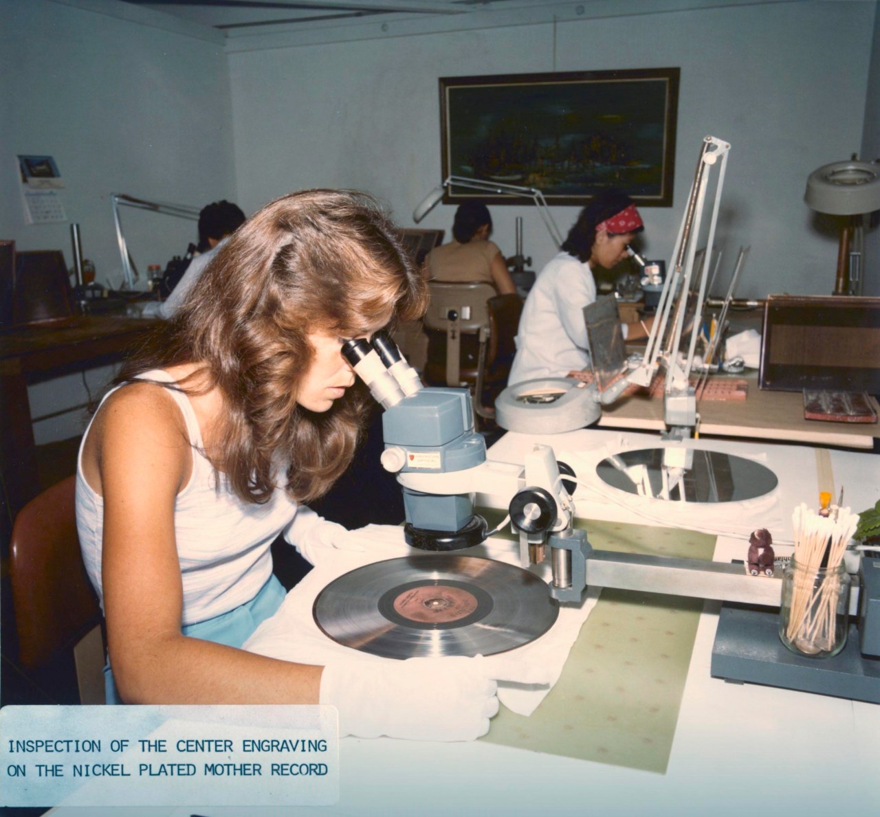 A worker wearing white gloves examines a silver mother record through a microscope at a crowded workbench.