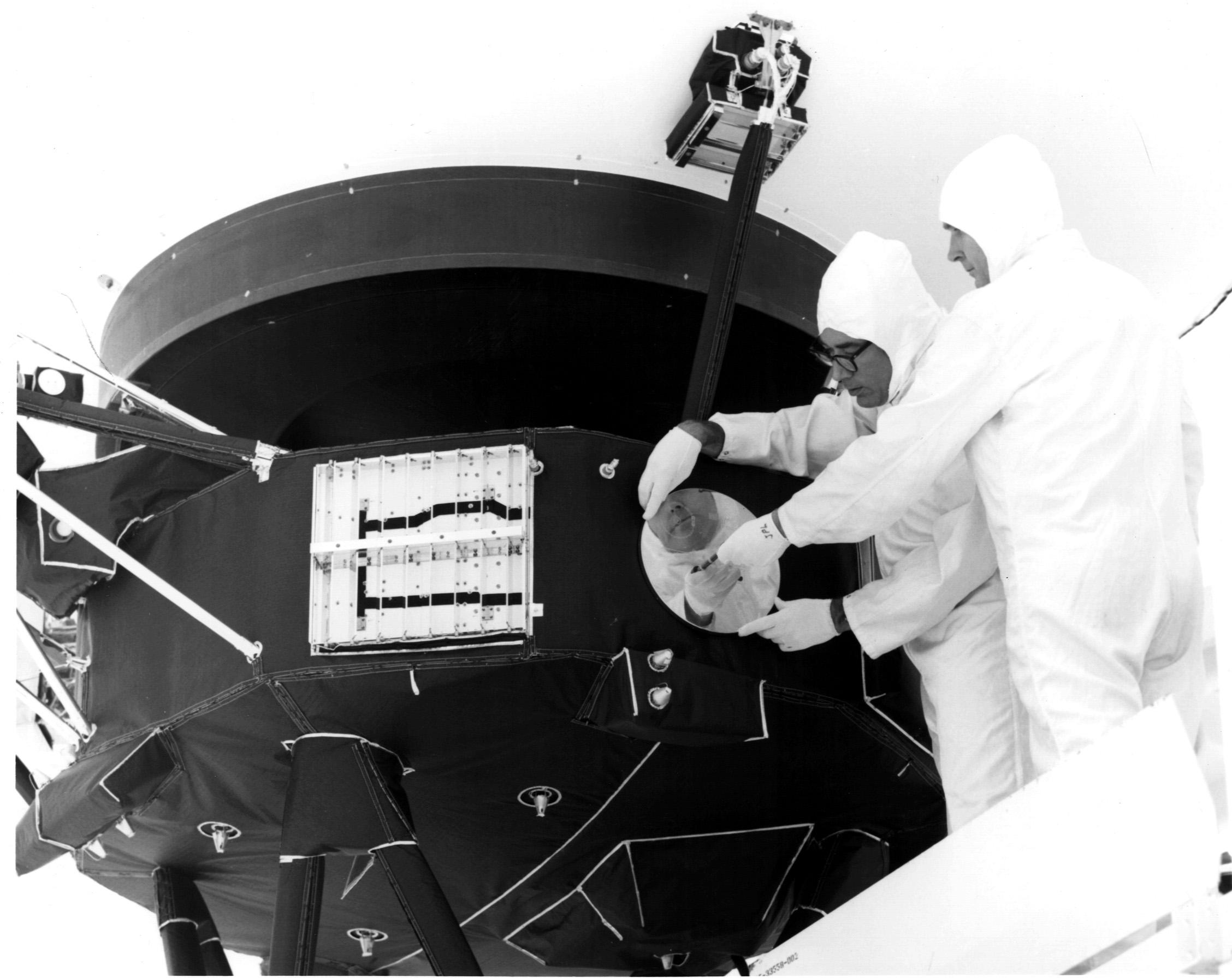 Two technicians beside Voyager 1 during the installation of its Golden Record package in 1977; the spacecraft’s large dish extends above them.